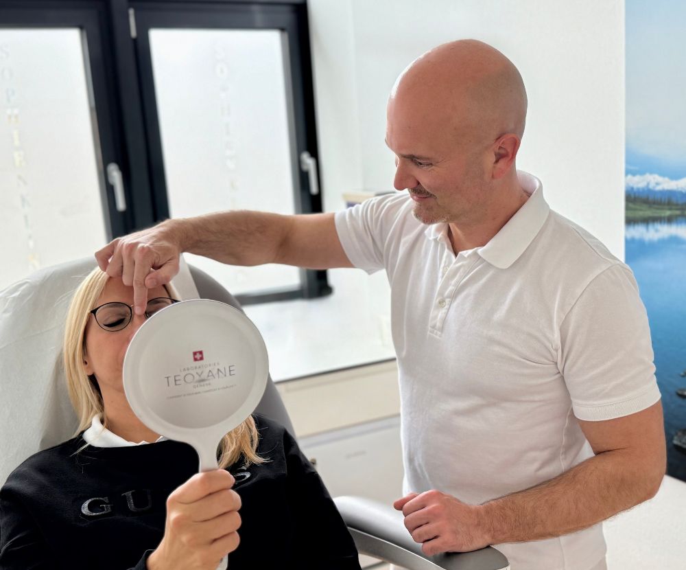 Dr. med. Gregor F. Fuchshuber demonstriert einer Patientin vor einem Spiegel, wie das Ergebnis einer Nasenkorrektur aussieht. Sie hält den Spiegel, während er ihr das Ergebnis zeigt.