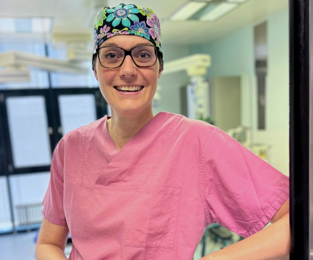 Dr. med. Sabrina Hoormann in pinkem OP-Outfit mit bunter Haube, lächelt vor der Klinik und bereitet sich auf eine Brustvergrößerung.