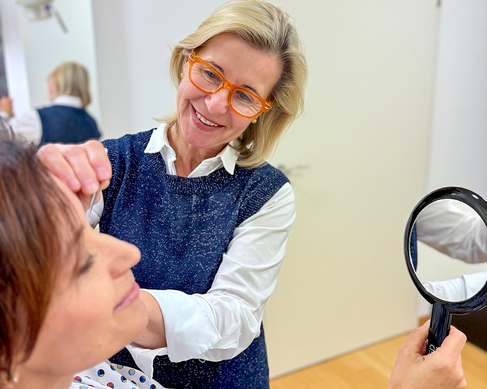 Dr. Dr. med. Susanne Herrmann-Frühwald untersucht bei einer persönlichen Beratung das Gesicht einer Patientin, die sich ein Facelifting in der Sophienklinik Stuttgart zeigen lässt. Die Patientin hält dabei einen Spiegel in der Hand, während die Expertin freundlich lächelnd die Gesichtspartien beurteilt.