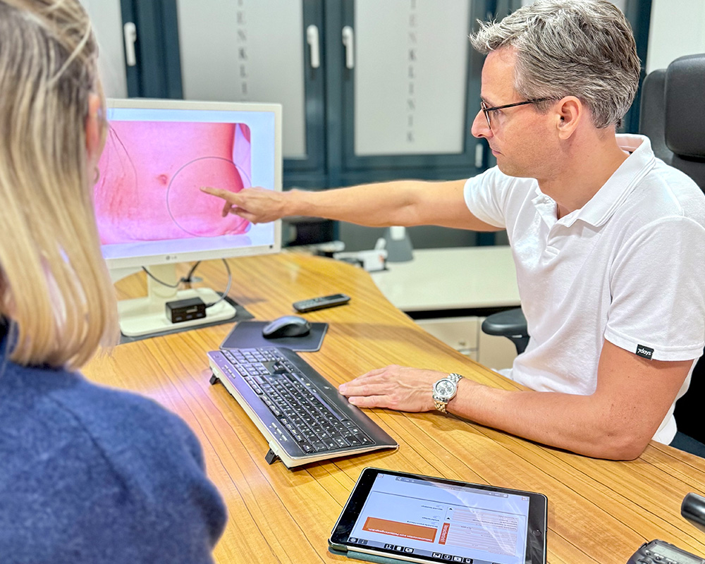 Dr. med. Konstantin Feise zeigt einer Patientin am Bildschirm eine vergrößerte Aufnahme eines Haut- und Gewebebereichs, um die Planung einer Fettabsaugung zu erläutern. Auf dem Schreibtisch liegen ergänzend Unterlagen und ein Tablet für die weitere Beratung.