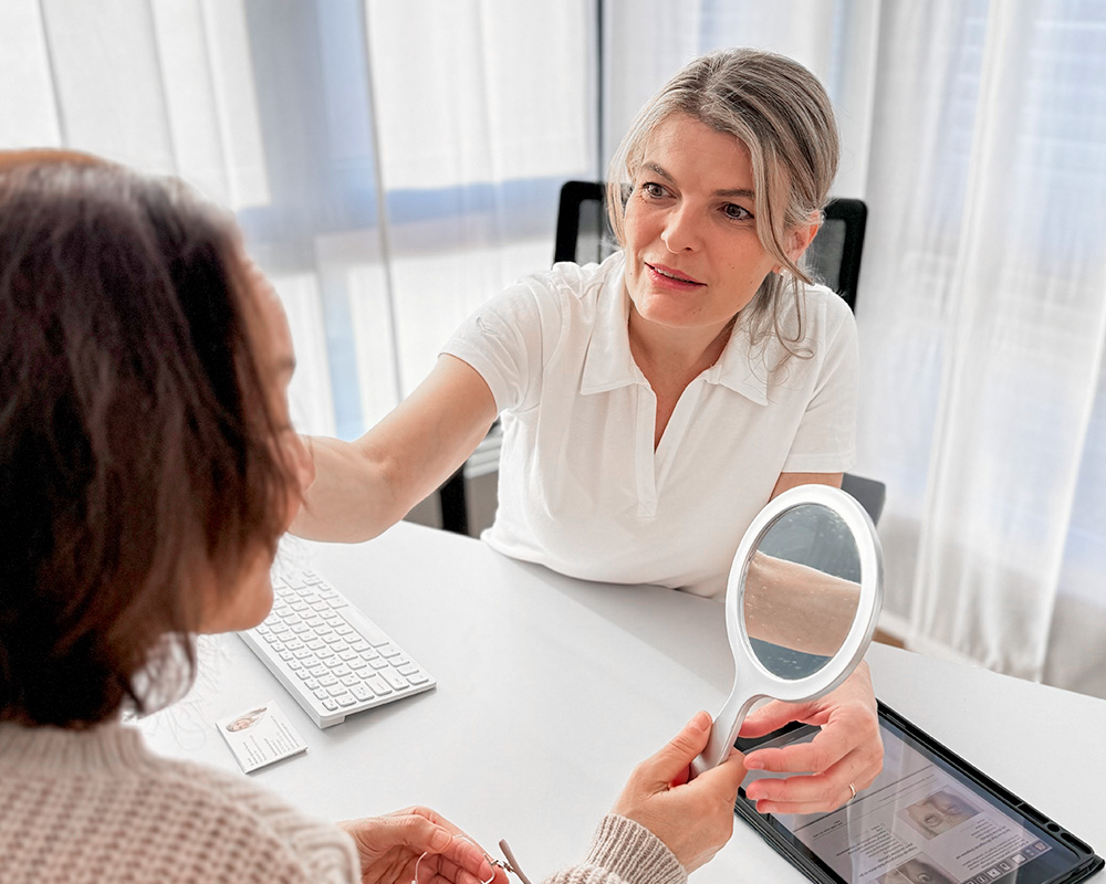 Dr. med. Mariann Schrick-Thomas betrachtet die Gesichtspartien einer Patientin und bespricht mögliche Behandlungsschritte. Die Patientin hält einen Spiegel, um die Empfehlungen direkt im eigenen Gesicht nachzuvollziehen.