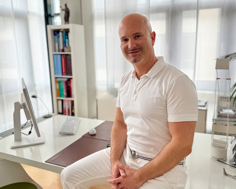 Dr. med. Gregor F. Fuchshuber lächelt im Büro der Sophienklinik und sitzt entspannt auf einem Stuhl. Im Hintergrund sind Bücherregale und ein Arbeitsplatz sichtbar.